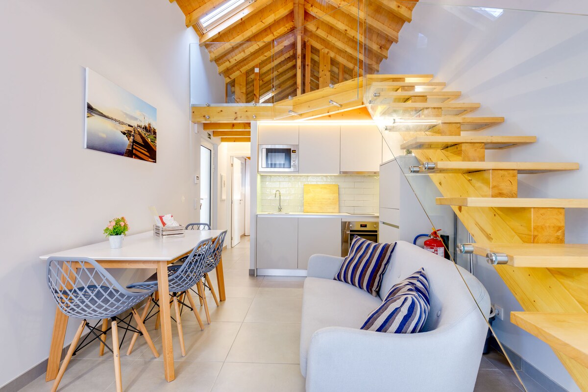 An open living area reveals a comfortable gray sofa with striped cushions, complemented by a modern dining table surrounded by designer chairs. A stylish wooden staircase leads to the upper level, and light fills the space through large windows.