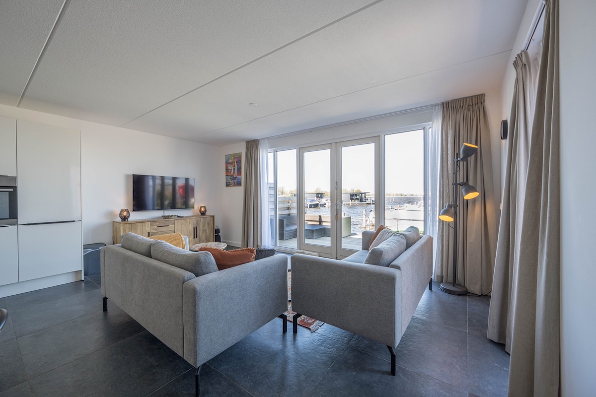 A bright living room features a comfortable seating area with two grey sofas arranged around a coffee table. Large glass doors open to a sunny terrace that showcases views of the harbor. Light-colored curtains frame the windows, complementing the overall design.