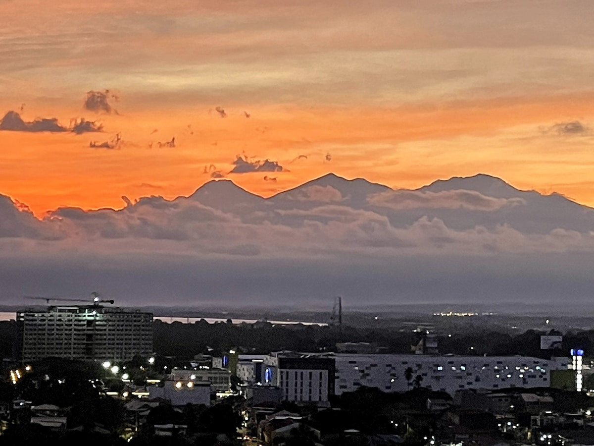 A striking view of Mt. Apo during sunset, showcasing various layers of color in the sky. The silhouette of the mountains stands out against the vibrant horizon, while city lights below create a contrast with the serene natural backdrop.