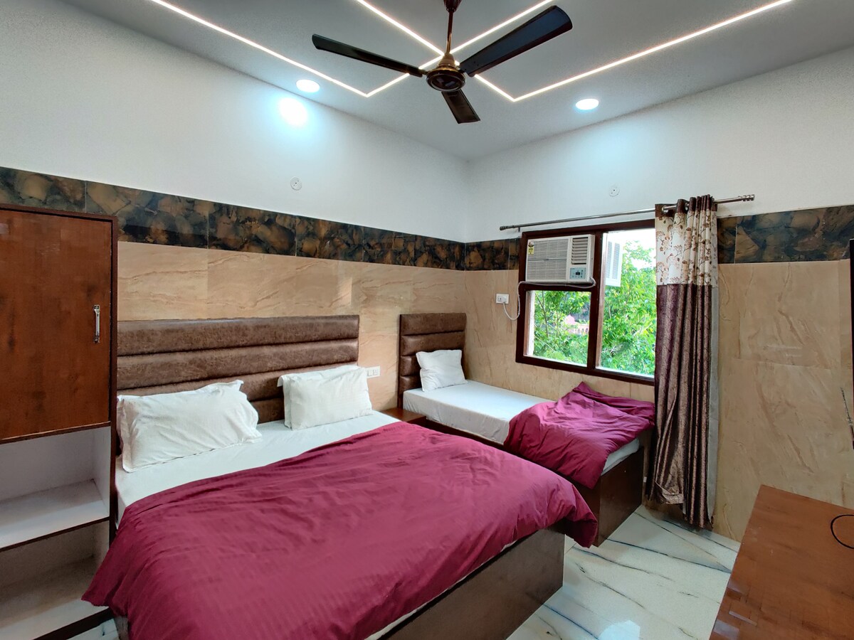 A comfortable sleeping area features a spacious bed with a deep red blanket. A second single bed is situated beside it, and an air conditioning unit is mounted above a window. Soft lighting is provided by recessed ceiling lights, complementing the room's neutral tones.