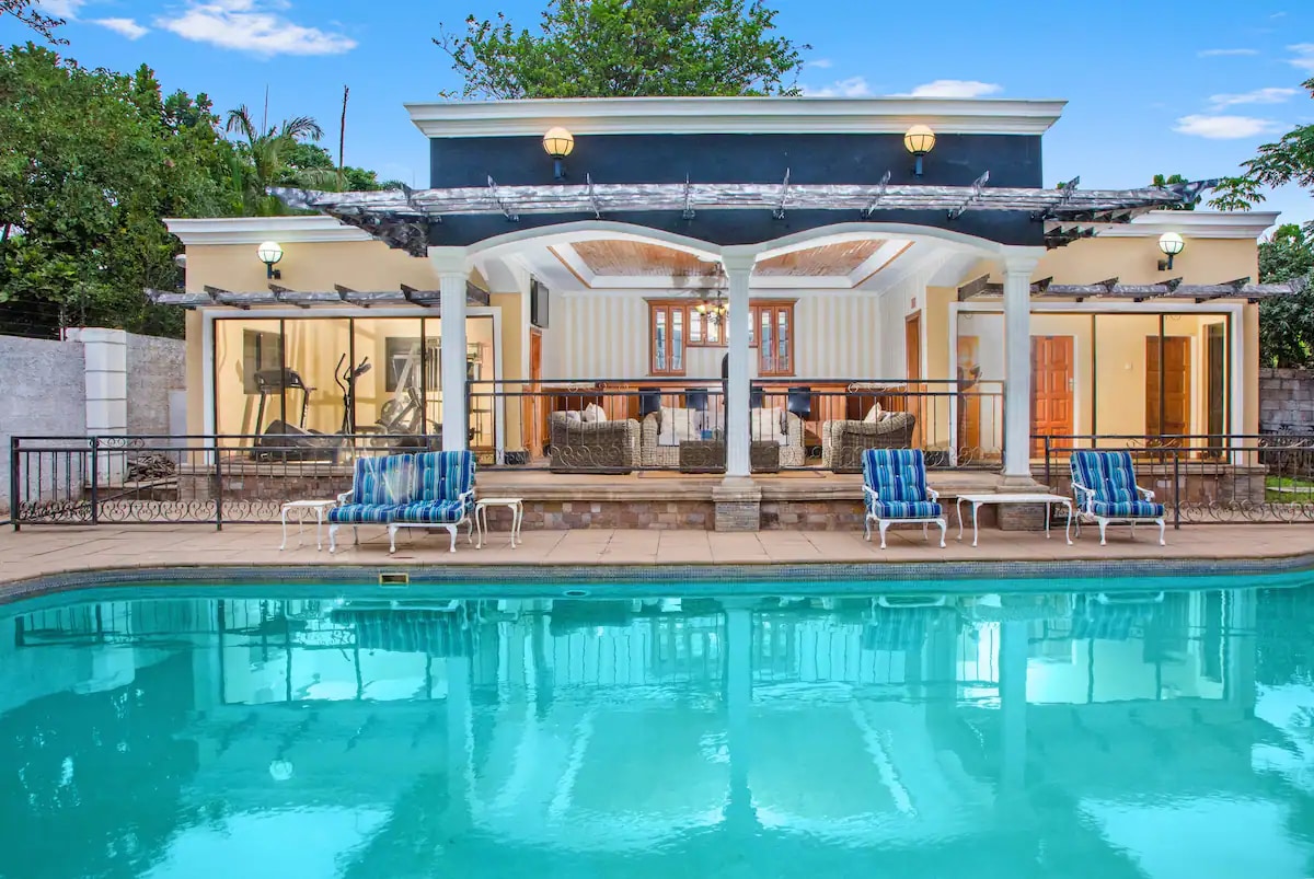 A well-maintained outdoor pool area is framed by a charming patio showcasing a covered seating area. Lounge chairs are positioned alongside the clear water, reflecting the blue sky. Abundant greenery is visible in the background, enhancing the tranquil environment.