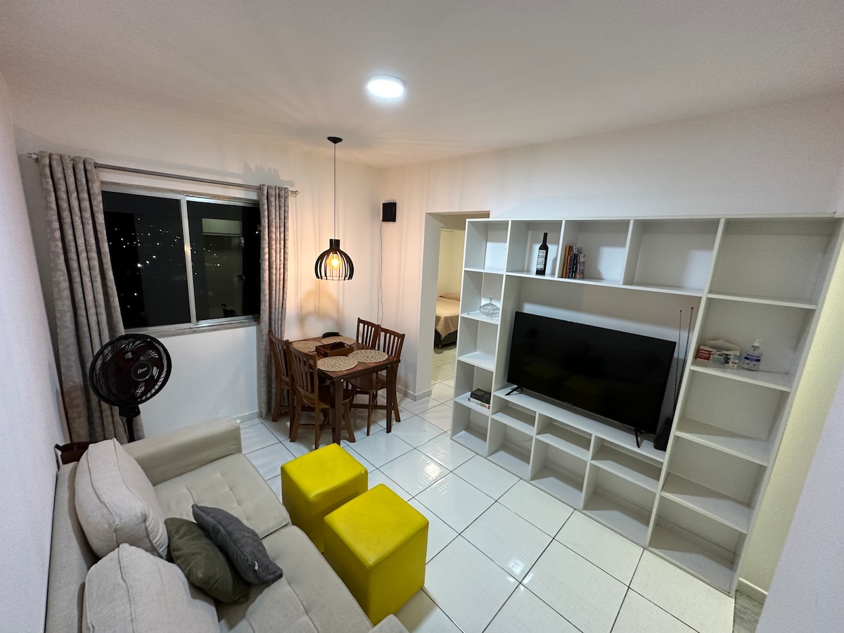 A living area features a sofa, two yellow stools, and a small dining table with wooden chairs. A wall-mounted entertainment unit holds a television, while a window provides natural light. Soft curtains frame the window, creating a bright and airy atmosphere.