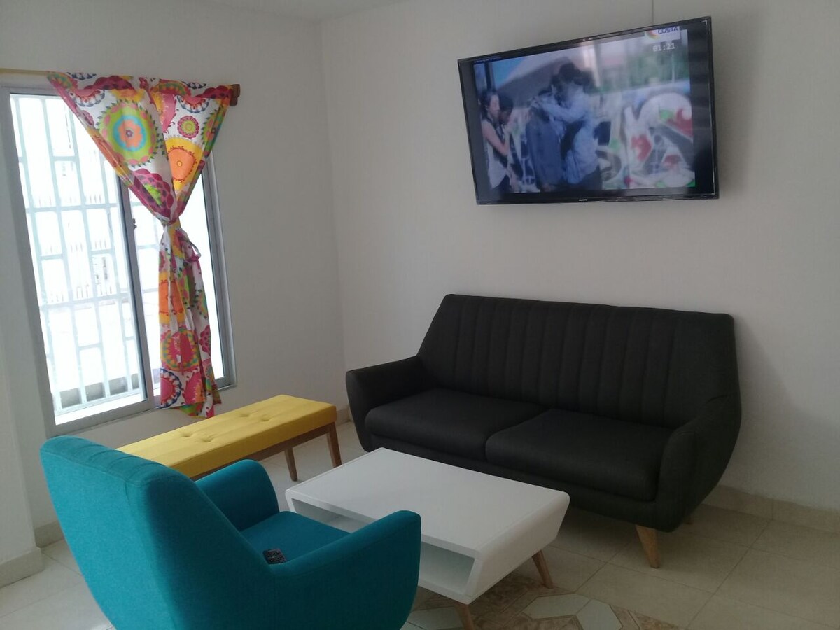 A cozy living area includes a black sofa and a colorful blue chair, complemented by a white coffee table. A bright yellow accent table adds a splash of color, while a large television is mounted on the wall. Natural light enters through a window adorned with vibrant curtains.