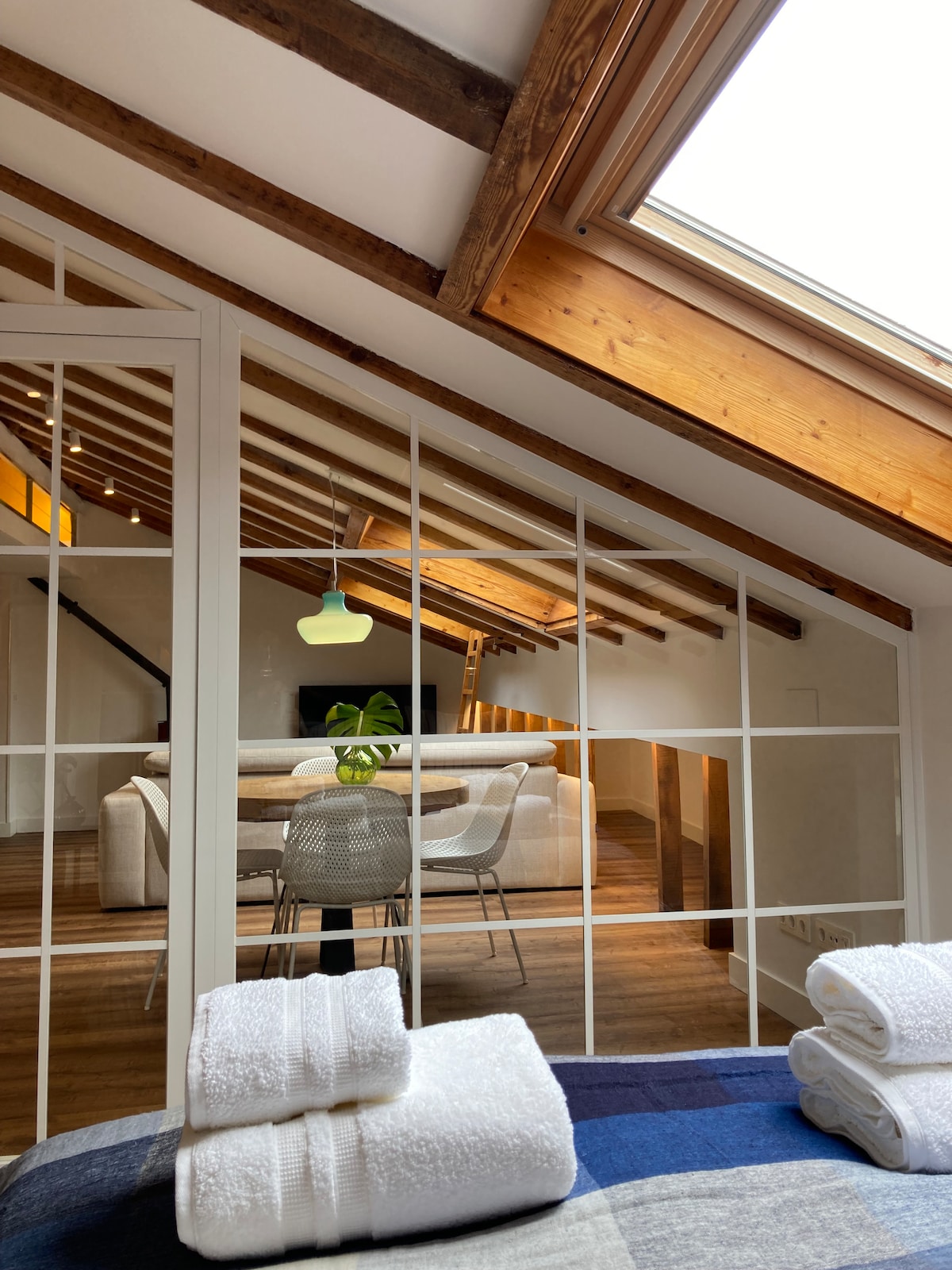 A spacious attic interior is visible through large glass doors, showcasing a well-lit living area with a comfortable sofa and dining table. Natural light illuminates the wood-beamed ceiling, while fresh towels are neatly arranged on a bed in the foreground.