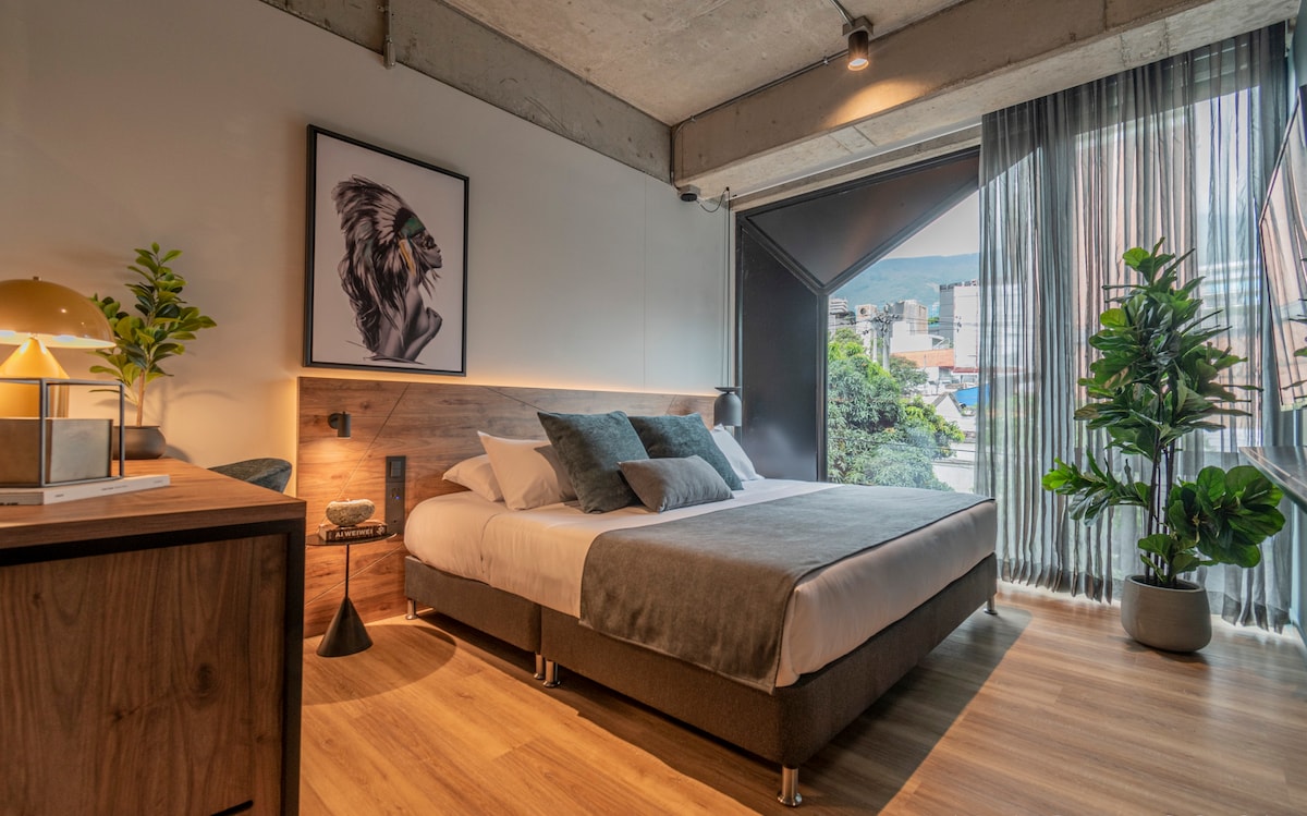 A modern bedroom features a spacious bed dressed in neutral tones, complemented by a large window adorned with sheer curtains. A piece of artwork decorates the wall above the bed, while potted plants and a bedside lamp add a touch of greenery and warmth to the space.