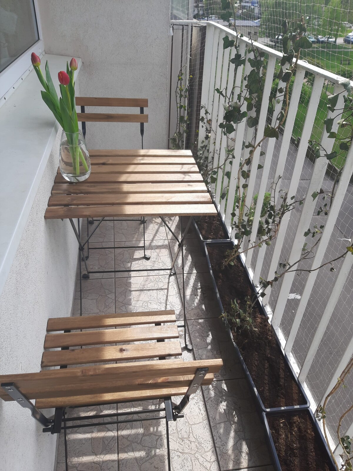 A small balcony featuring a wooden table and two folding chairs, positioned against a wall. Fresh tulips are displayed in a glass vase on the table. The floor is tiled, and planter boxes line the edges with growing plants, contributing to a green space.