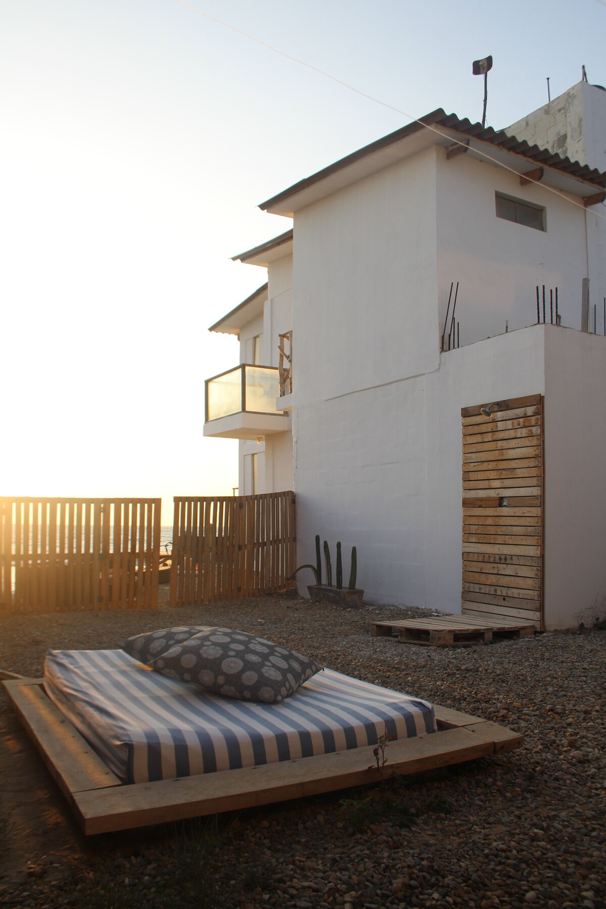 A sunlit exterior view of a two-story duplex showcases a simple wooden patio with a cushioned outdoor bed. The surrounding space features a gravel surface, with a wooden fence and cacti in the background, creating a serene and inviting atmosphere.
