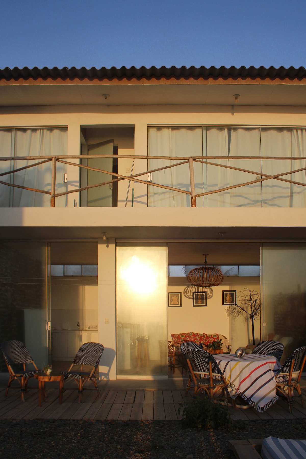 An outdoor seating area is visible, featuring multiple wooden chairs and a dining table. The second floor presents a balcony with glass doors, allowing natural light to fill the space. The warm light of the setting sun reflects off the glass, creating a serene atmosphere.
