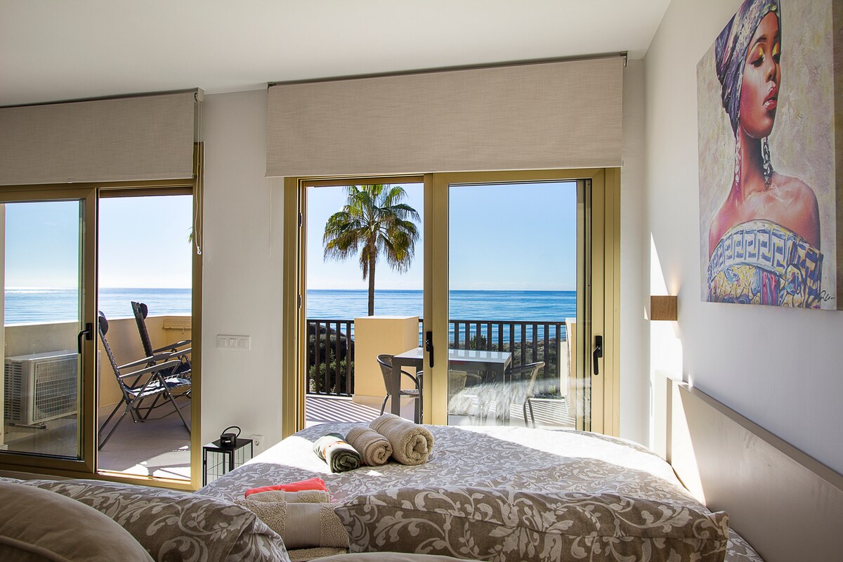 The spacious studio features large glass doors opening to a private terrace, providing unobstructed views of the sea. A cozy bed is adorned with neatly arranged towels, while the serene decor and natural light create a calming ambiance.
