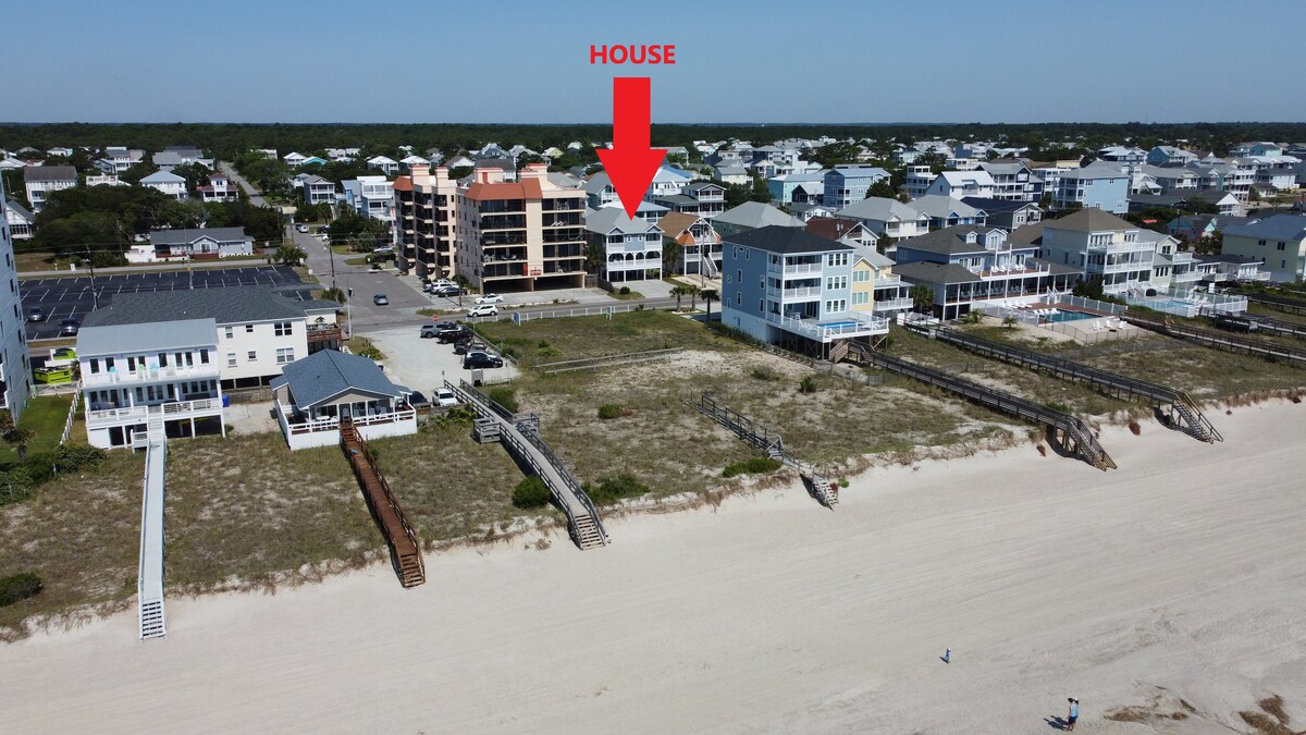 An aerial view of the beach house, prominently marked, displaying its proximity to the coast. Surrounding structures show a mix of residential homes and rental properties, with sandy beach access visible. The beach stretches alongside the shore, defining the landscape.