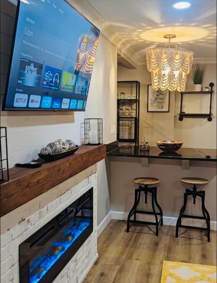 A modern living area features a wall-mounted TV above a sleek electric fireplace with blue illumination. A stylish chandelier hangs from the ceiling, illuminating a small bar area with two stools and a bowl, set against a neutral wall.
