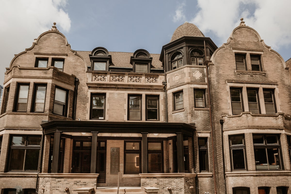 The exterior of a historic building features ornate architectural details and prominent gabled roofs. The façade is composed of bricks in a light tone, with large windows and an inviting front porch. The surrounding area is open to the sky, complemented by scattered clouds.