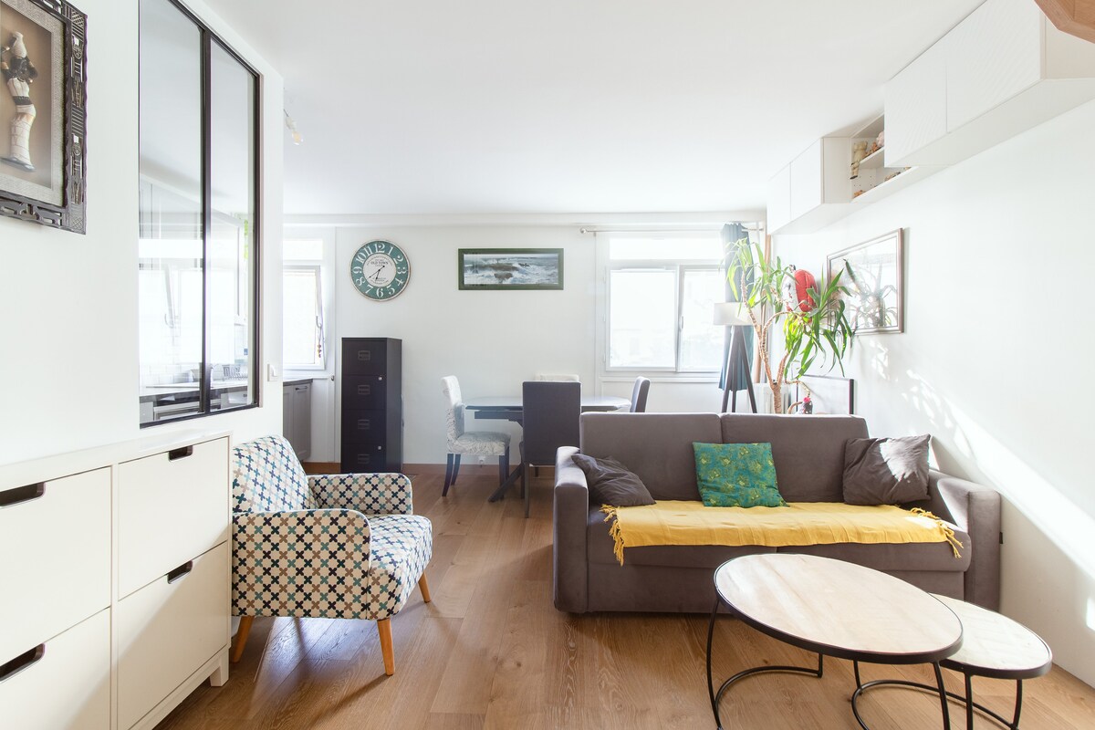 A bright living space features a comfortable gray sofa and an accent chair adorned with a patterned fabric. A light wooden coffee table sits in front, while a dining area with a small table and chairs is visible in the background. Natural light fills the room through several windows.