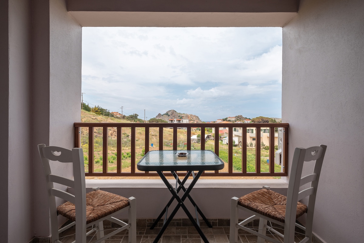 A balcony is shown with two white chairs and a small table positioned in front of a wide, open window. The view reveals rolling hills and a distant landscape, while soft natural light enhances the scene.