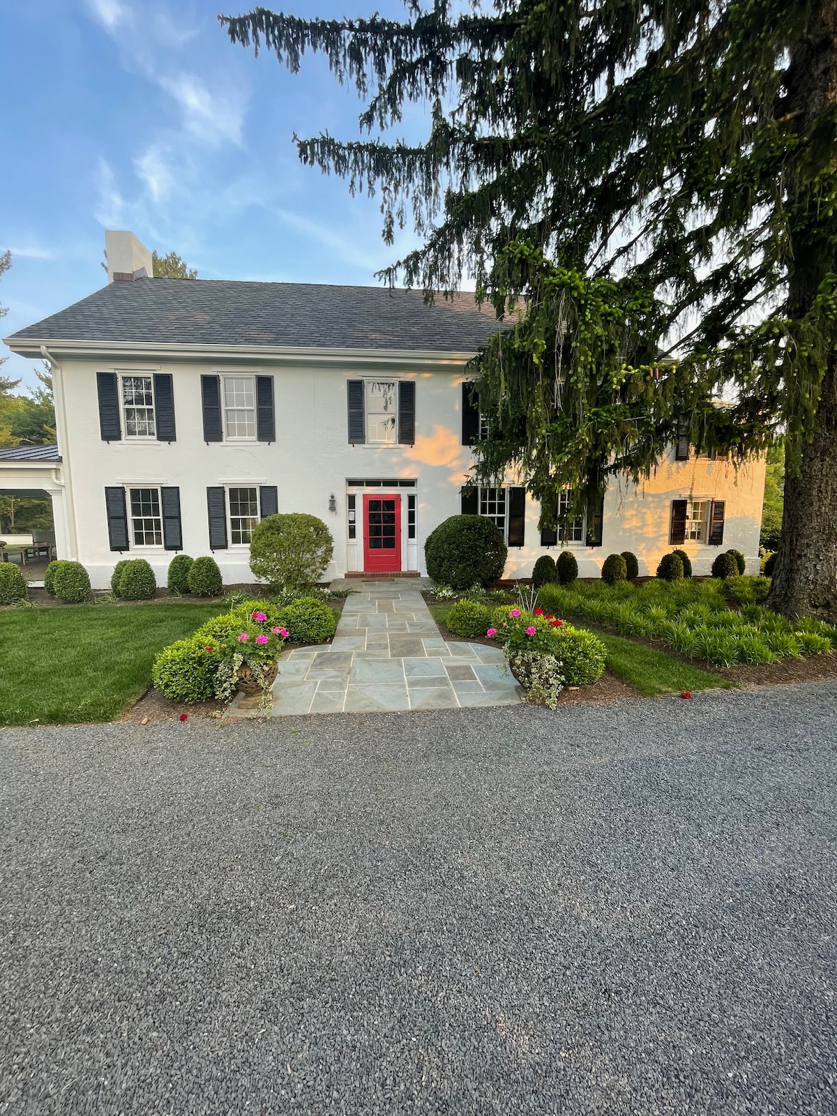 A classic two-story farmhouse is depicted, featuring a white exterior and black shutters. A vibrant red front door is centered within the facade. Manicured green shrubs and colorful flowerbeds flank the stone pathway leading to the entrance, while tall trees provide shade nearby.