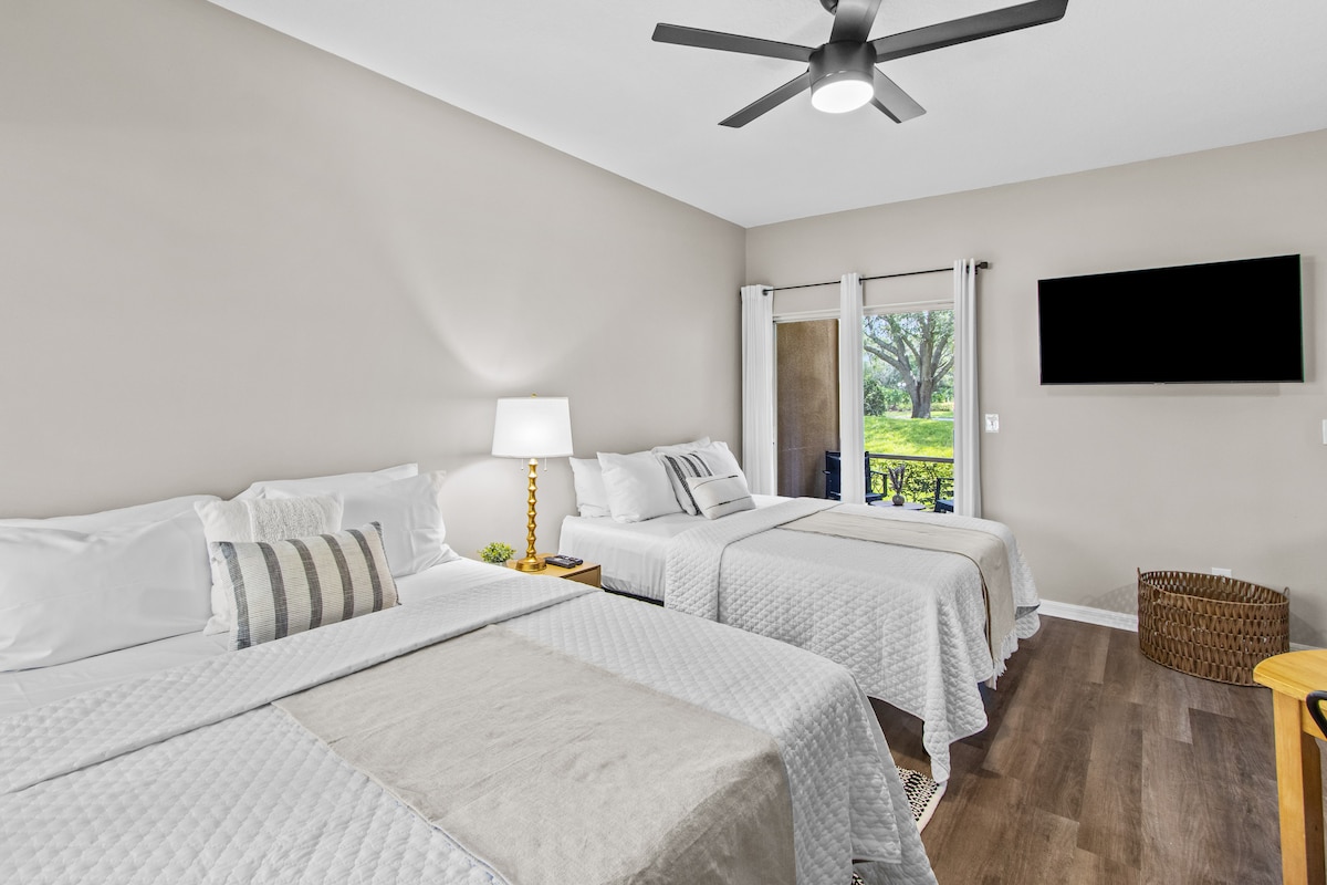 A bright bedroom is equipped with two spacious beds, each topped with white linens and decorative pillows. A subtle beige wall enhances the serene atmosphere. A large window offers views of the outdoors, while a ceiling fan provides airflow. A flat-screen TV is mounted on the wall.