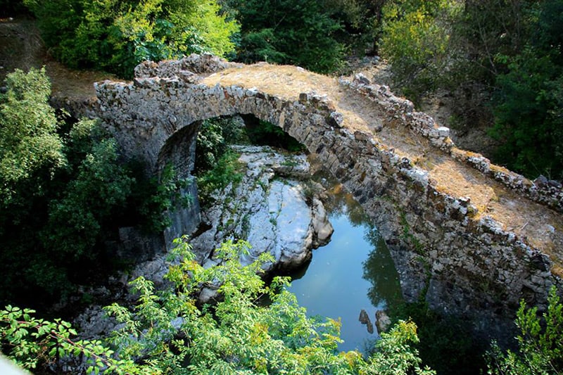 An ancient stone bridge arches gracefully over a tranquil body of water, surrounded by lush greenery. The structure showcases weathered stones, blending harmoniously with the natural landscape. Reflections in the water enhance the serene atmosphere of the setting.