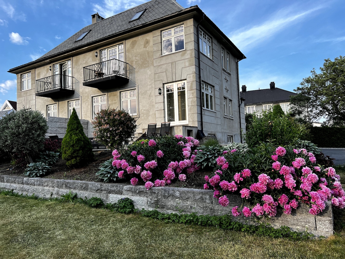 A two-story building is set amidst a landscaped garden, featuring vibrant pink flowers and lush greenery. Balconies are visible on the upper level, while a neat stone flower bed lines the front. A grassy area extends in front of the structure, enhancing the serene environment.