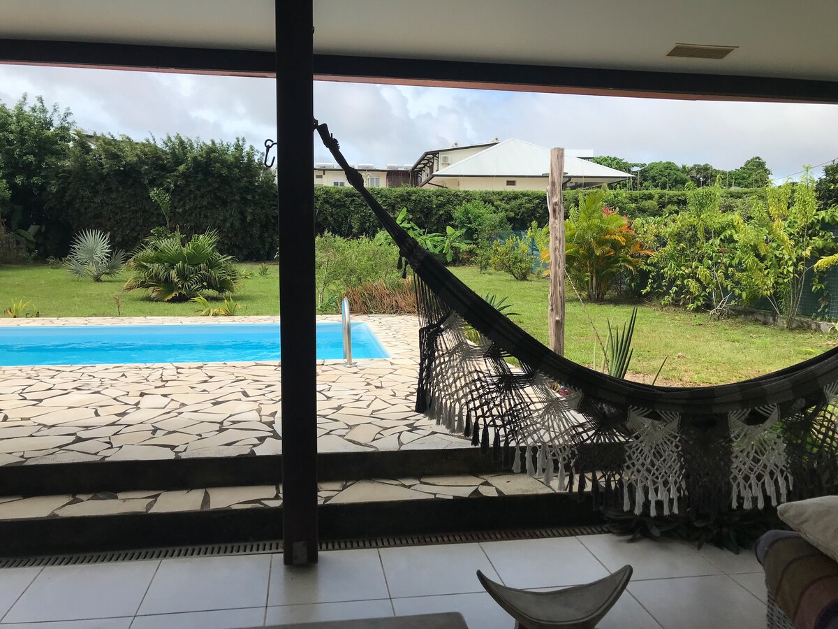 A hammock is suspended under a covered terrace, providing a view of the lush garden and a blue swimming pool. The landscape includes various plants and trees, with a clear sky above, creating a tranquil setting for relaxation.