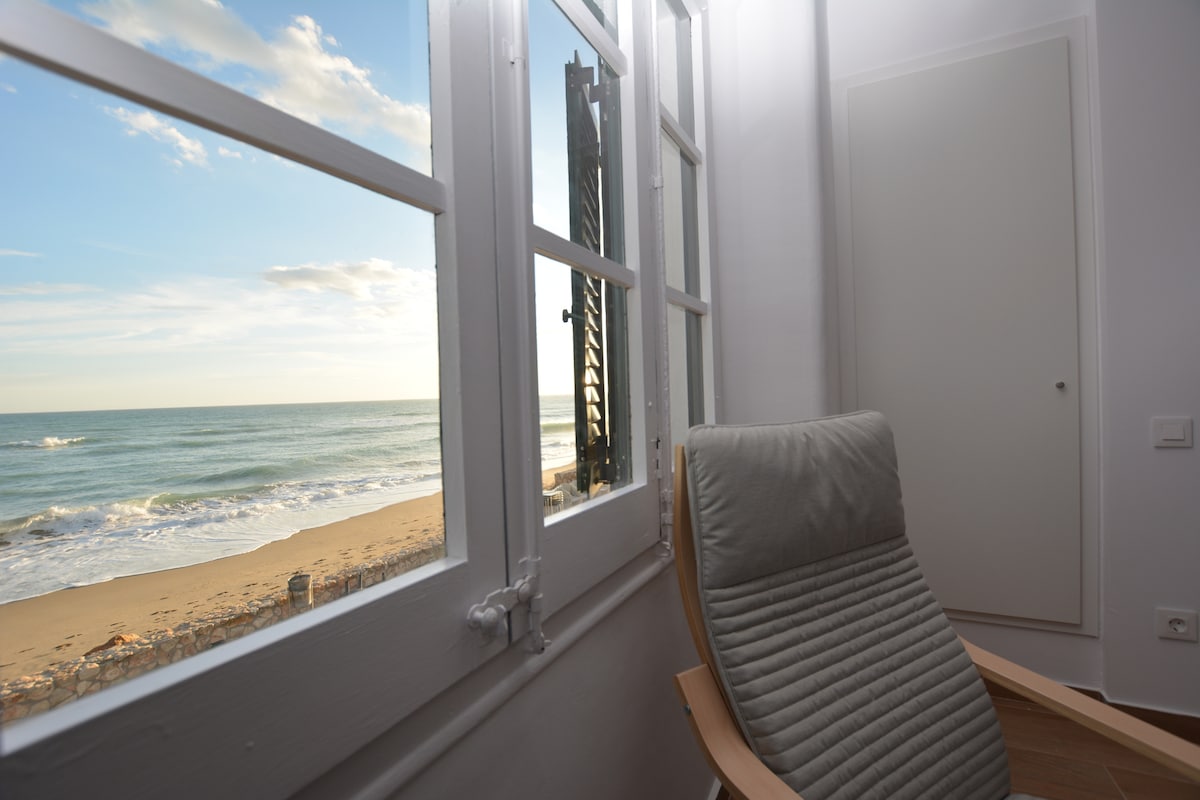 A well-lit seating area features a comfortable chair positioned by a large window. Outside, views of the sandy beach and gentle ocean waves are visible, with a clear sky above. The window allows natural light to fill the room, enhancing the coastal ambiance.