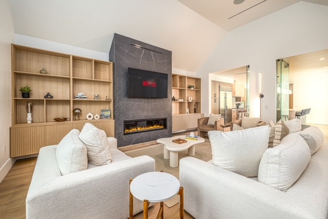 A modern living area features comfortable, light-colored sofas arranged around a central coffee table. A sleek fireplace adds warmth, while a large TV is mounted on the wall. Built-in shelves display decorative items, providing an organized and inviting space for relaxation.