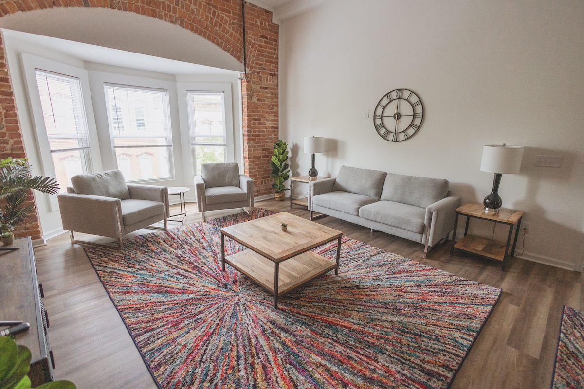 A spacious living area features a vibrant area rug with multicolored patterns, complementing the exposed brick walls. Two comfortable armchairs and a light grey sofa are arranged around a wooden coffee table. Large windows allow natural light to illuminate the space, creating a welcoming atmosphere.
