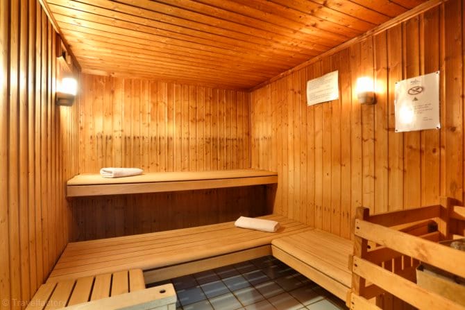 The sauna features warm wooden paneling and two tiered benches for relaxation. Soft lighting enhances the serene atmosphere. A small wooden structure holds towels and additional items, providing a comfortable environment for rejuvenation.