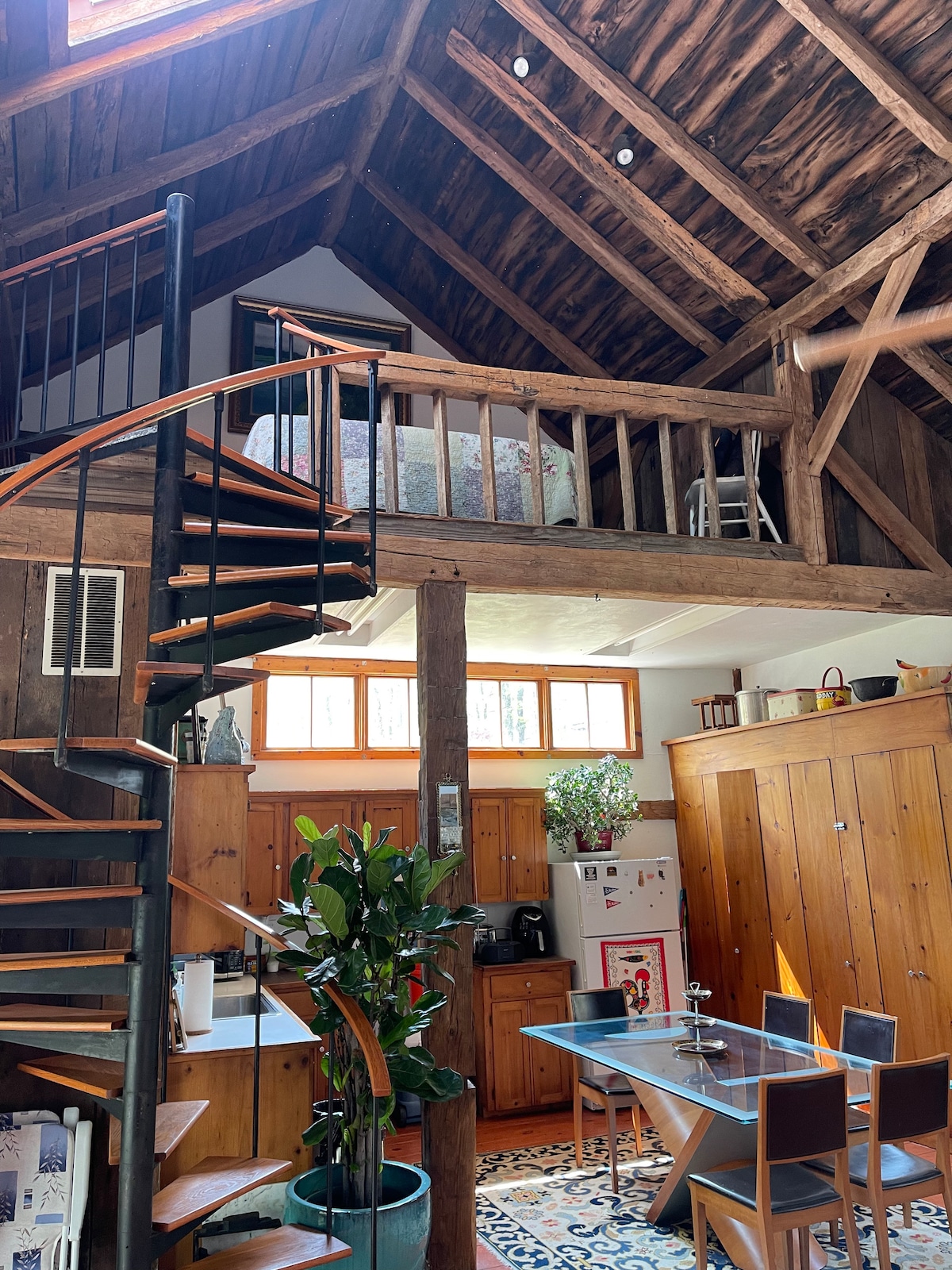 A spiral staircase leads to a sleeping loft, featuring a spacious bed and wooden accents. Natural light streams through multiple windows, illuminating the rustic wooden walls. A dining table sits below, surrounded by chairs, with a kitchenette visible in the background.