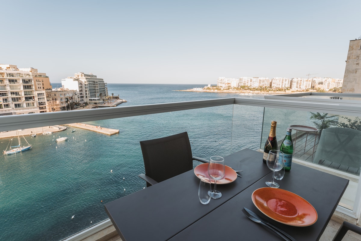 A balcony table is set for dining, featuring two plates and glasses beside bottles of wine and sparkling water. The view includes calm waters of Spinola Bay and surrounding buildings, with a yacht visible in the distance under a clear sky.