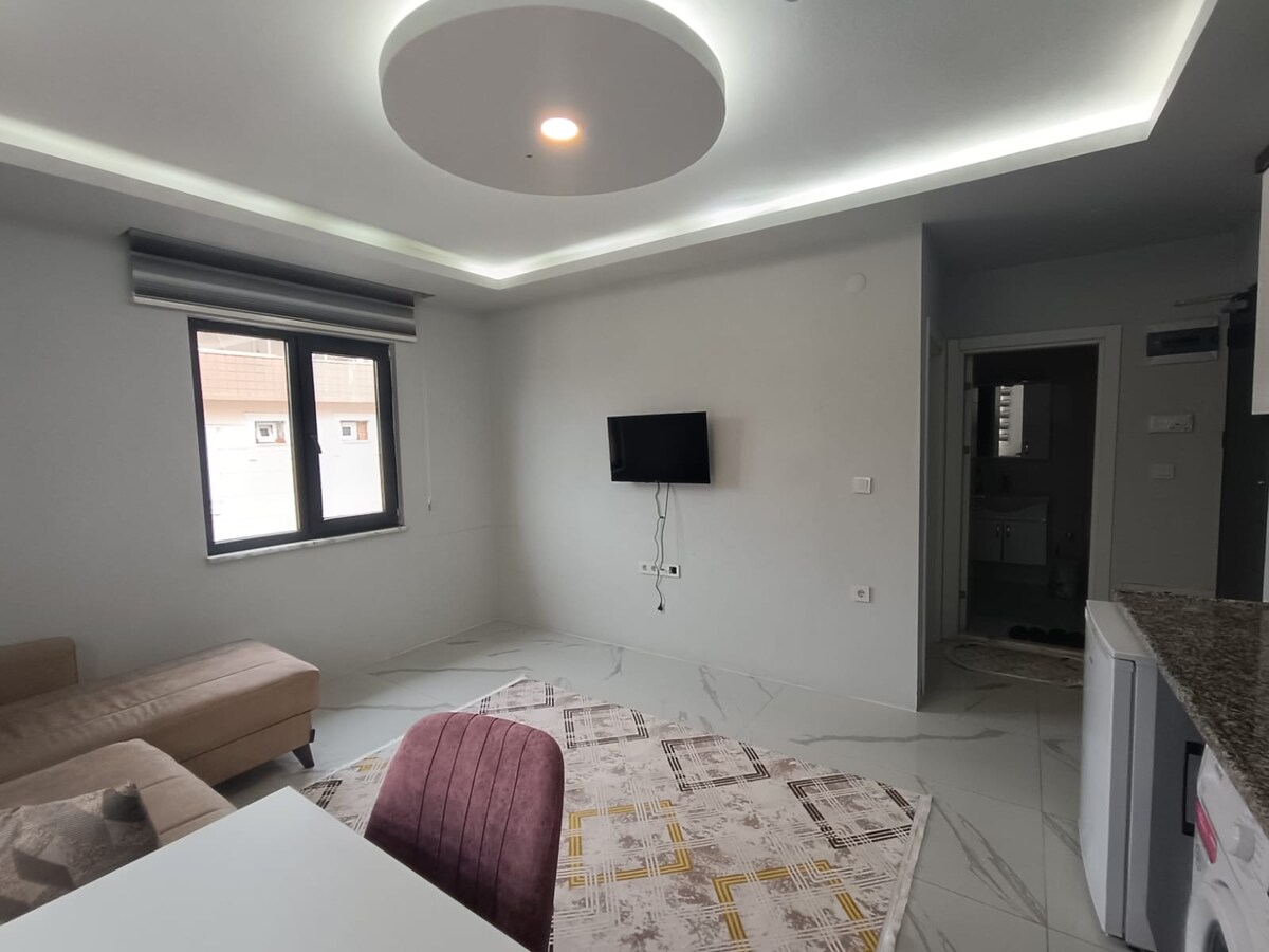 The living area is characterized by a modern design, featuring a beige sofa and a dining table. A flat-screen television is mounted on the wall, while soft LED lighting illuminates the ceiling. Large windows provide natural light and a view of the outdoors.