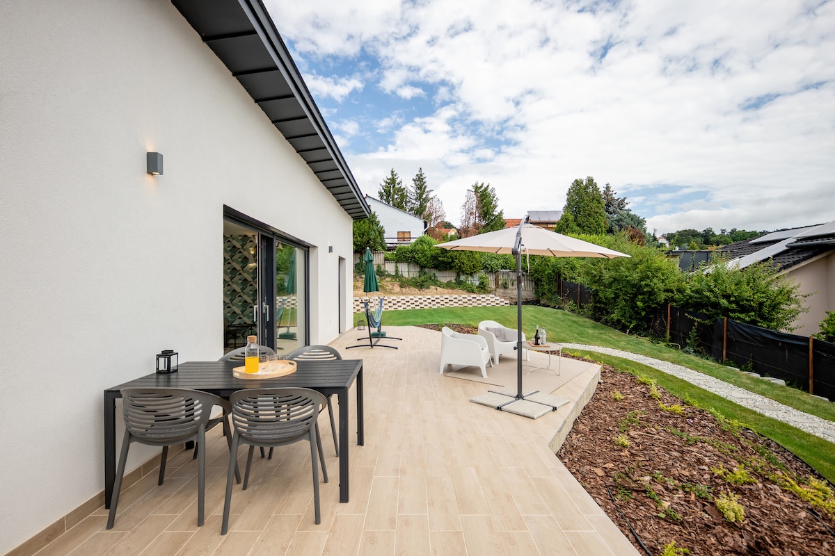 An outdoor terrace is displayed, featuring a dining table with four chairs and an umbrella providing shade. Sun loungers are positioned nearby, surrounded by green grass and landscaped areas. The sky is partly cloudy, contributing to a serene atmosphere.