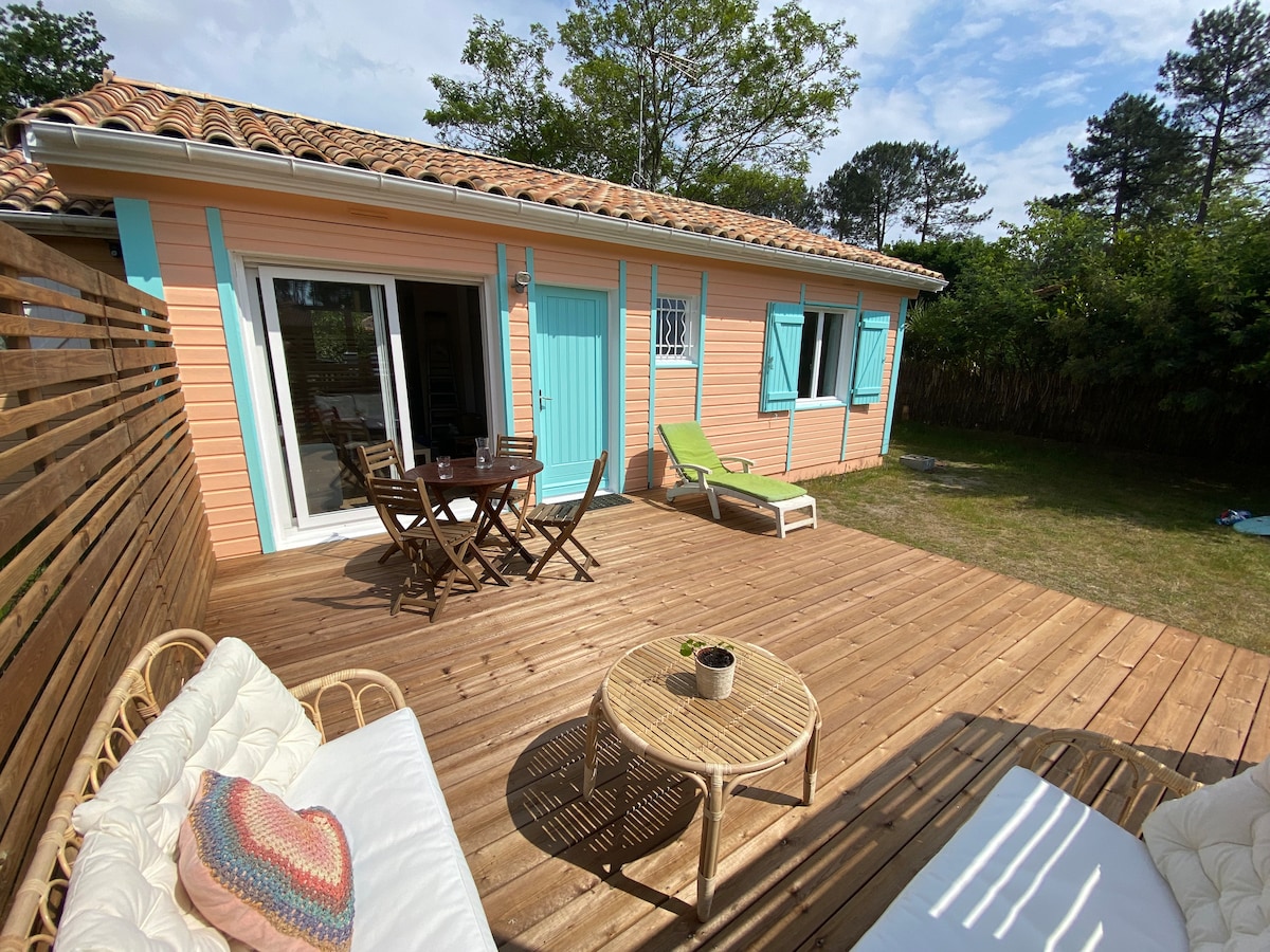 A wooden terrace is featured outside the house, furnished with a dining table and chairs, alongside two lounge chairs. The house, painted in soft colors, includes large glass doors leading to a garden area surrounded by greenery.
