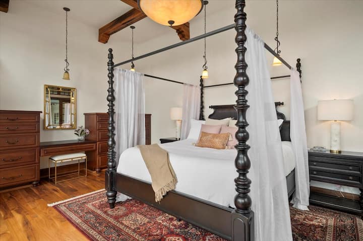 The south wing of the main floor master king bedroom, located off the main living room. A four-poster bed, floor-to-ceiling windows, a walk-in closet, a walk-out balcony with furniture, a primary bathroom with a double vanity and walk-in rain shower.