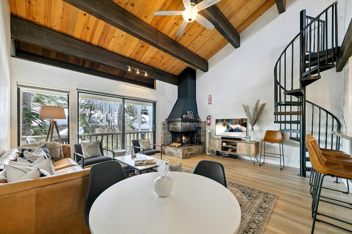 An open living area showcases a mix of comfortable seating and a sleek coffee table under a wooden ceiling with exposed beams. A stone fireplace serves as a focal point, while large windows offer views of the surrounding nature. A spiral staircase leads to the loft area.