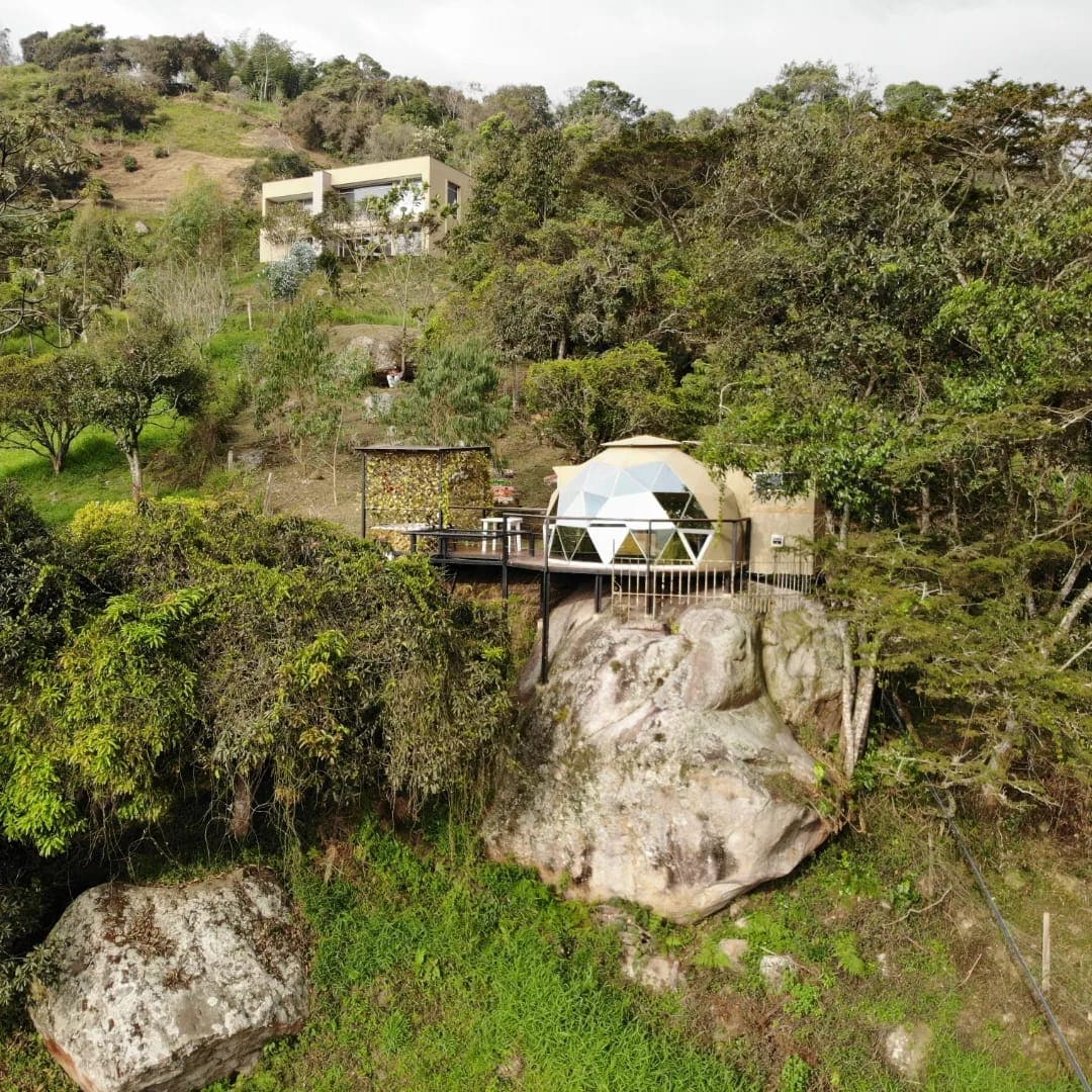 A geodesic dome is situated on a rocky outcrop, surrounded by lush greenery. Nearby structures blend with the natural landscape, while a spacious deck extends from the dome, offering scenic views of the hillside. The environment conveys a sense of tranquility and privacy.