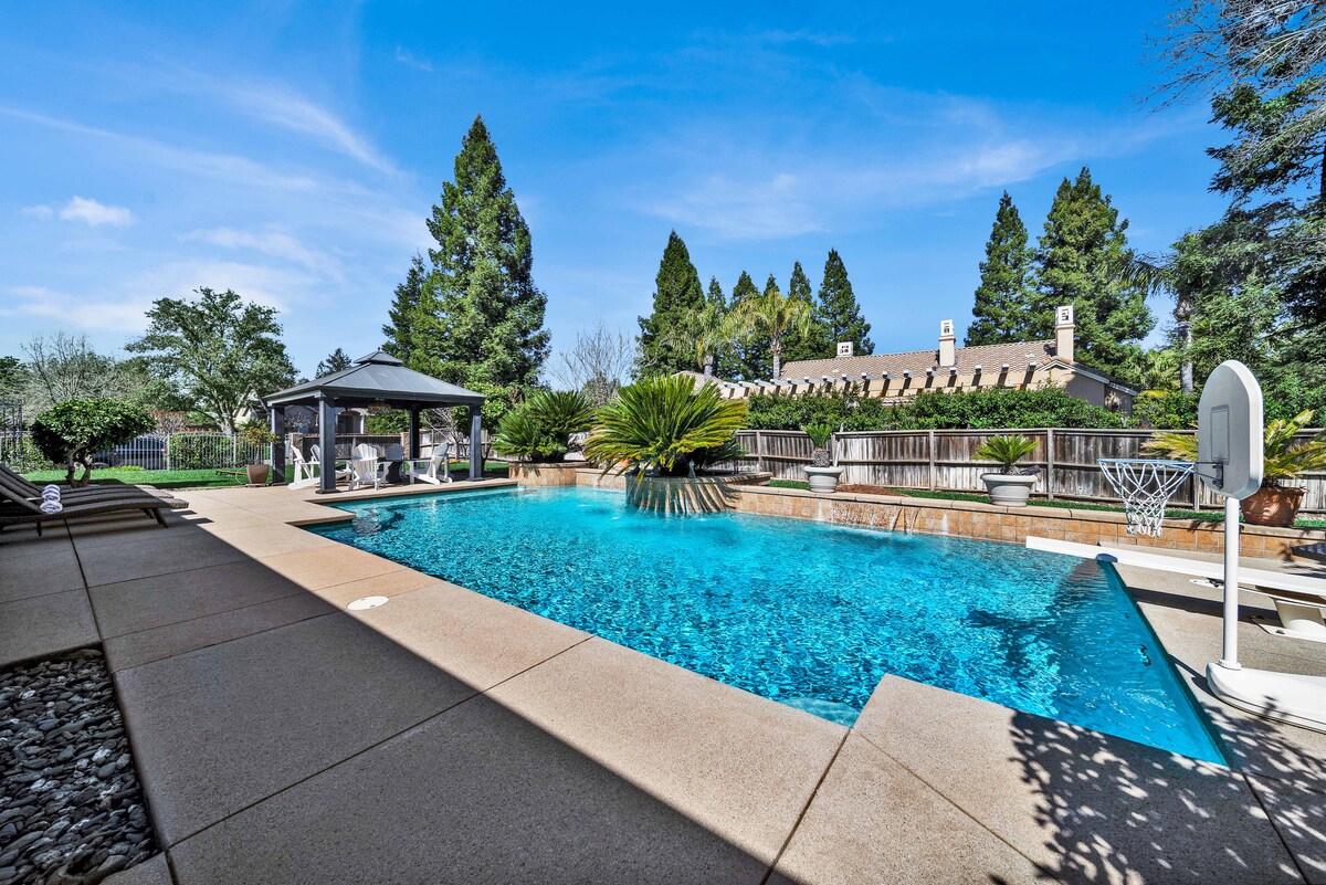 The backyard features a sparkling pool surrounded by a spacious patio and lounge chairs. A gazebo provides shaded seating, while tall trees and landscaped greenery create a serene atmosphere. The pool area is ideal for relaxation and outdoor activities.