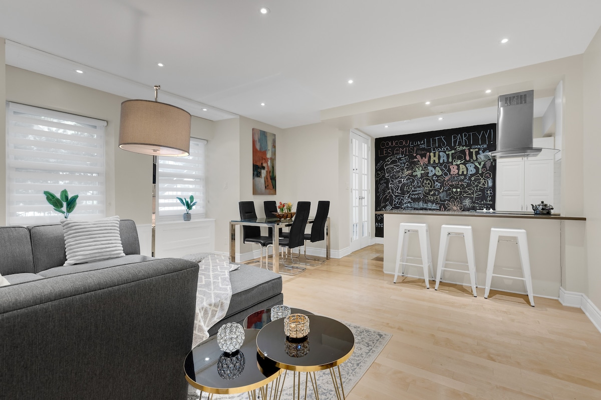 A modern living area features a comfortable gray sofa and a sleek coffee table with decorative elements. A dining area with stylish chairs is visible, while a chalkboard wall adds a playful touch to the kitchen space. Bright lighting enhances the open layout.