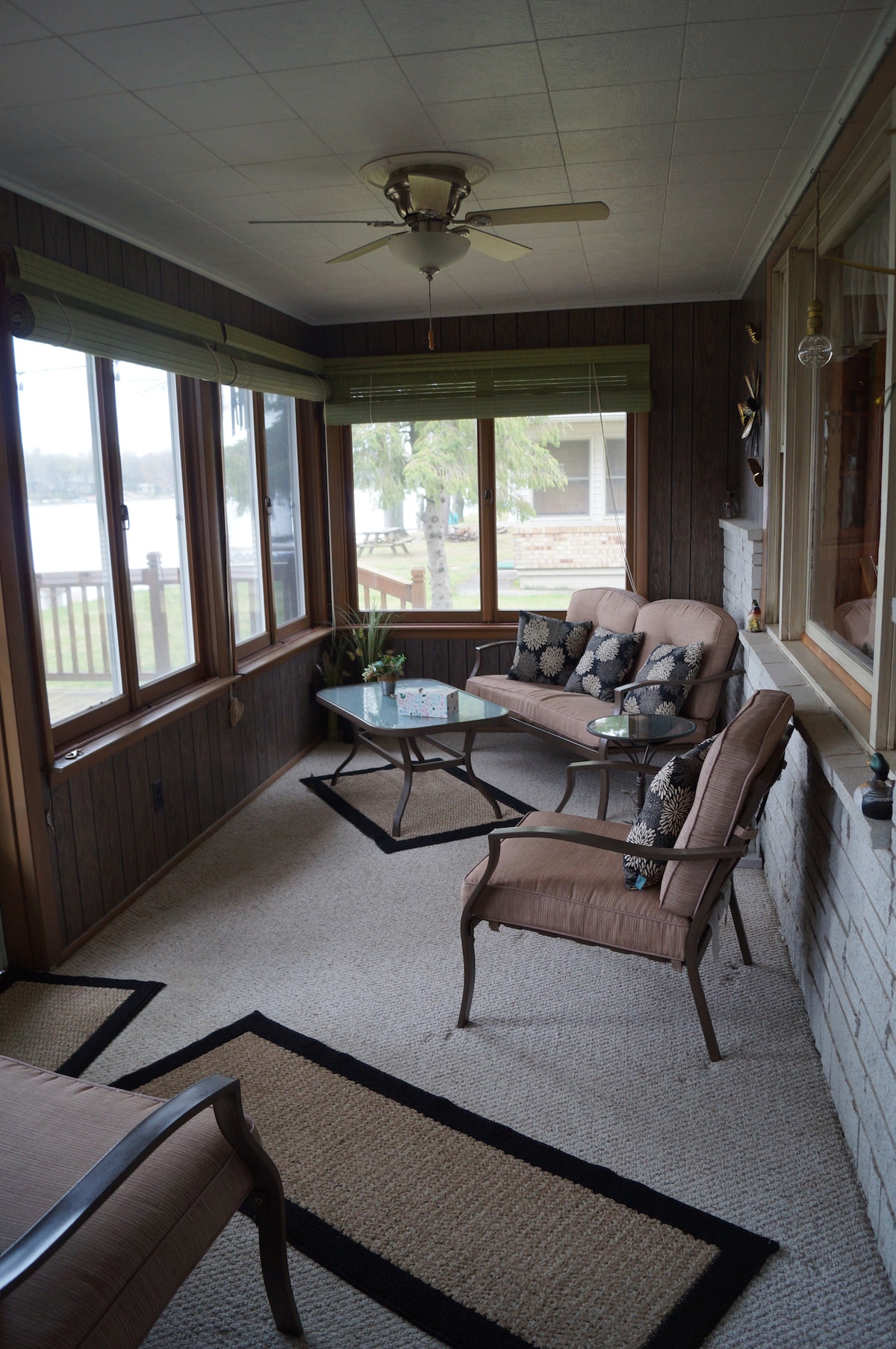 A bright three-season room features panoramic windows allowing natural light and views of the lake. Comfortable seating arrangements include two chairs and a sofa with decorative pillows, centered around a glass-top table. A ceiling fan provides a gentle breeze in the cozy space.