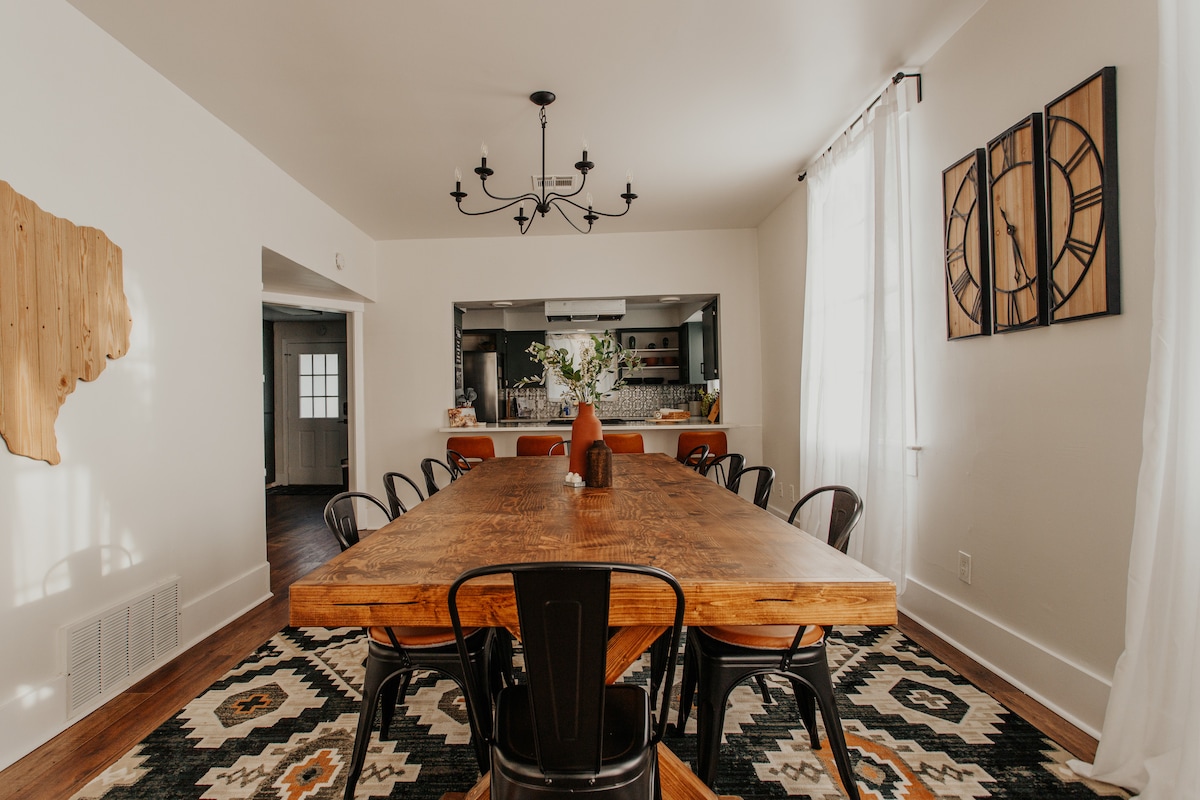 A spacious dining area features a large wooden table surrounded by black chairs. A pendant chandelier hangs overhead, while a patterned rug adds texture to the floor. Natural light flows through large windows adorned with sheer curtains, creating a warm and inviting space.