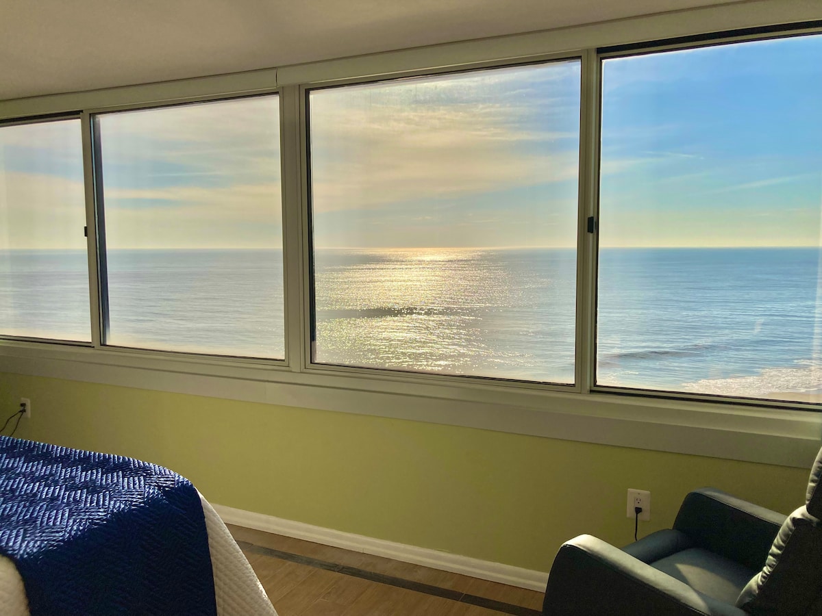 A panoramic view from the master bedroom showcases the expansive ocean, reflecting sunlight across the water's surface. Large windows allow natural light to illuminate the room, enhancing the serene atmosphere of the coastal environment.