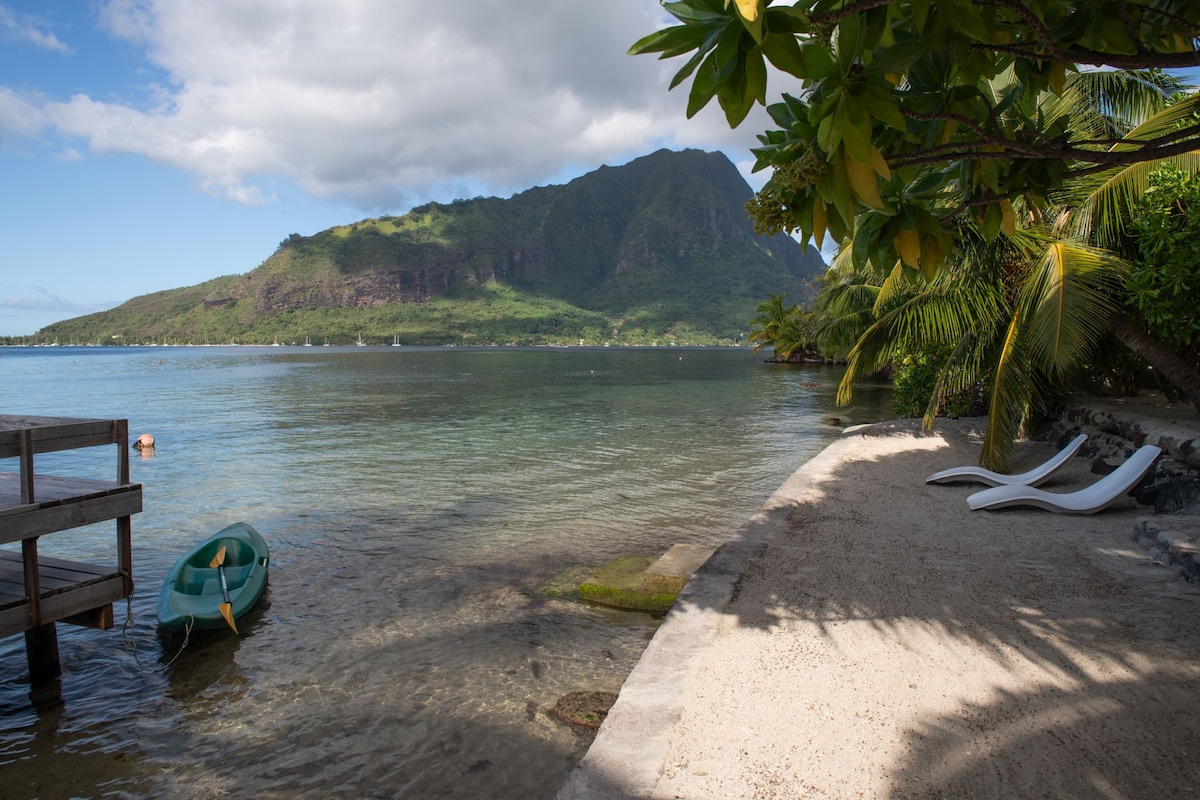 The image captures a serene beach scene with a clear view of a calm lagoon and a distant mountain. A kayak rests at the water's edge, alongside two sun loungers under overhanging tropical foliage. Soft sand leads to the gentle lapping of the water.