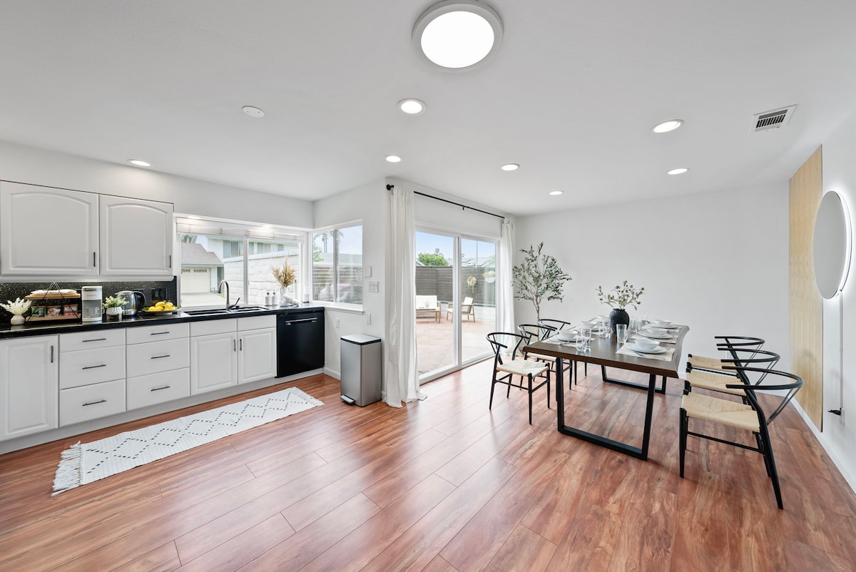 The dining area features a sleek wooden table set for six, complemented by black chairs. A large window allows natural light to flood the space, offering a view of the outdoor area. The kitchen, equipped with modern appliances, is visible in the background.