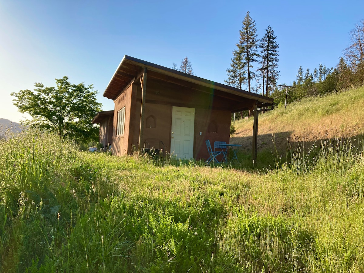 The earthen guest cottage is nestled among lush green grass, showcasing a quaint exterior with a porch and two blue chairs. Tall trees rise in the background under a clear blue sky, creating a serene setting in Southern Oregon.