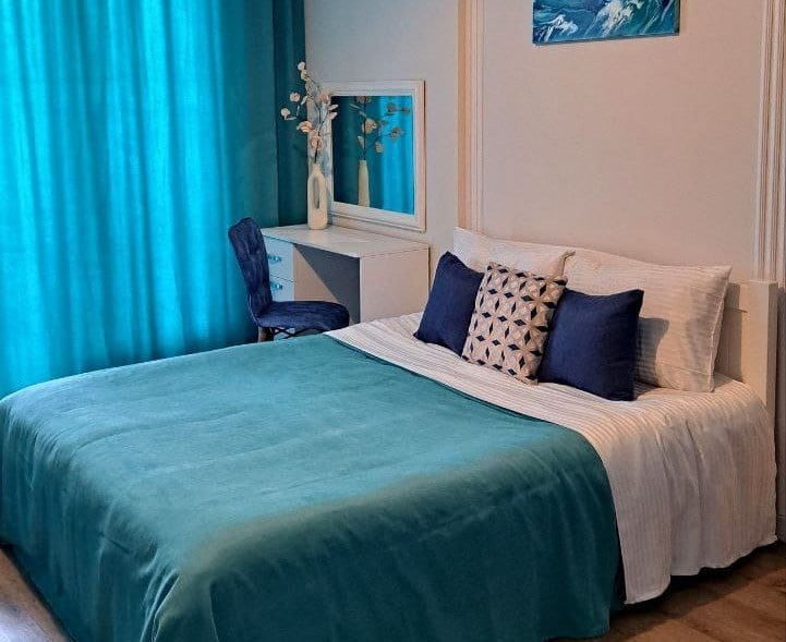 A spacious bed is draped in a soft teal blanket, complemented by decorative pillows in varying shades of blue. A mirrored vanity sits beside the bed, flanked by a chair in a matching hue and adorned with a vase of flowers. Floor-to-ceiling curtains in a similar teal provide a colorful backdrop.
