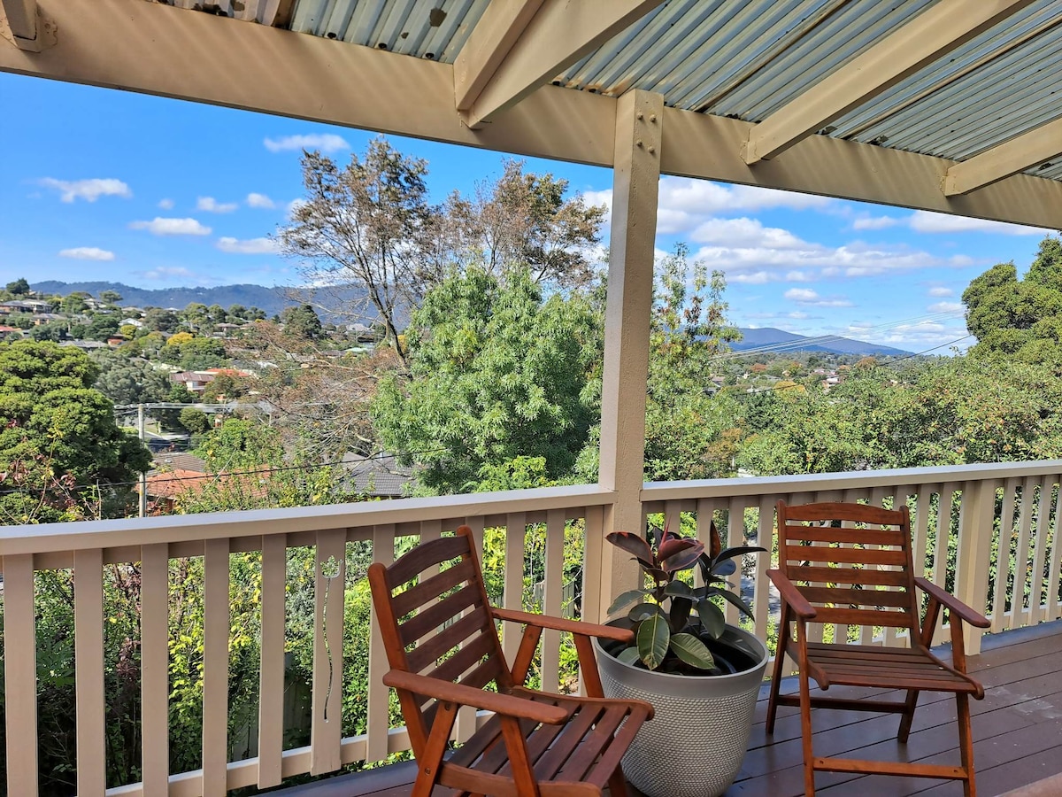 A wooden deck offers a comfortable space with two chairs positioned for enjoying sweeping views of the surrounding trees and mountains. Bright blue skies and scattered clouds create a serene backdrop, enhancing the tranquil setting.