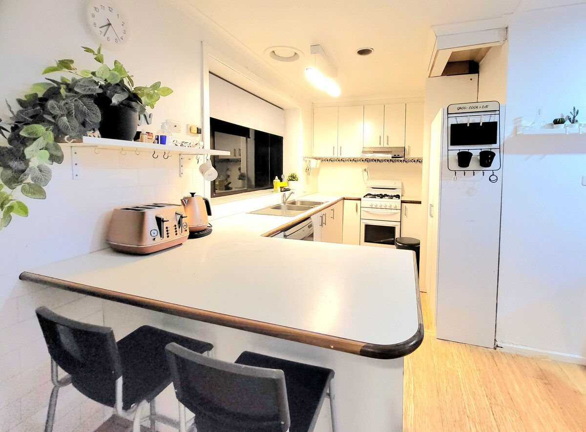 A bright full-size kitchen is illuminated by overhead lighting, featuring modern appliances including a stove and fridge. A spacious countertop extends into a breakfast bar with two black chairs. Natural light streams through a large window adorned with minimalist decor.