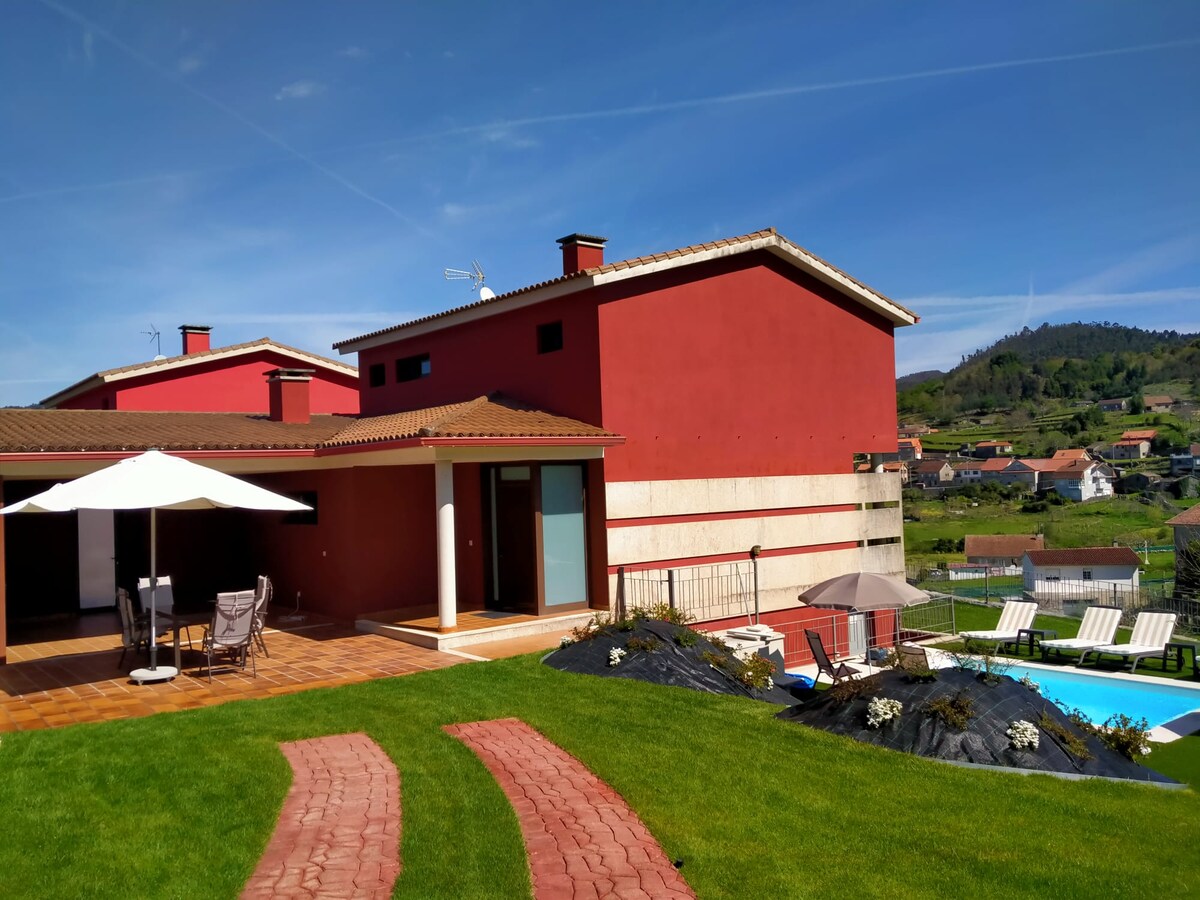 A spacious outdoor area is featured with well-maintained grass and stone pathways leading to a swimming pool. The villa, painted in a warm hue, is flanked by lounge chairs and an umbrella for shade, set against a backdrop of rolling green hills.