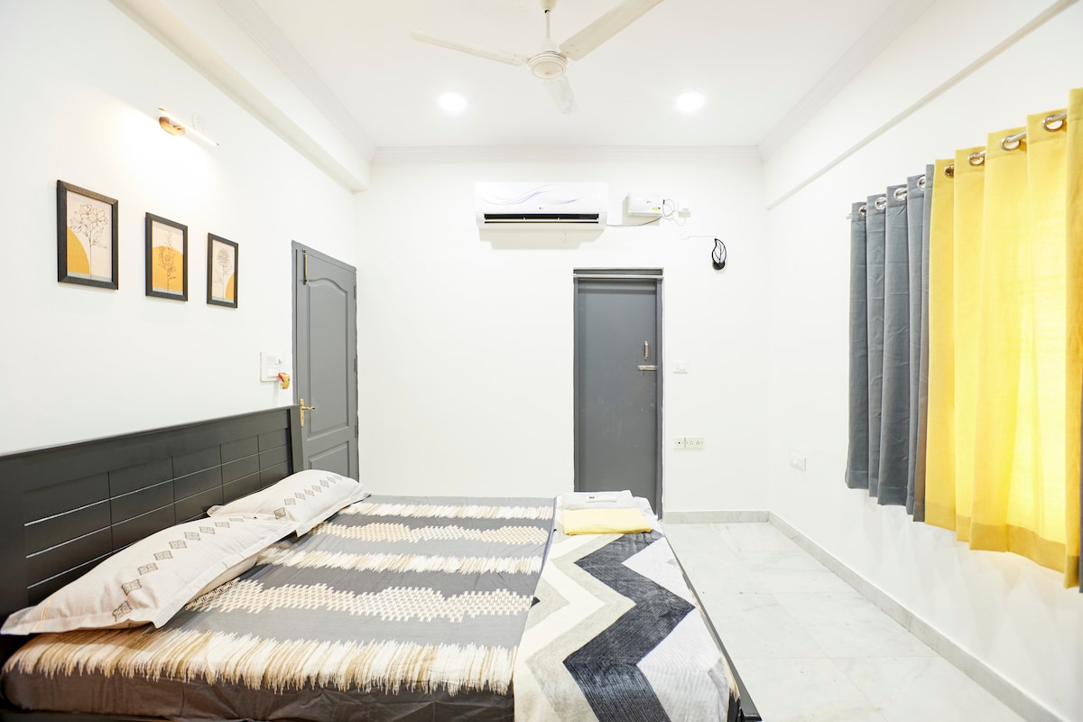 A well-lit, spacious room features a large bed with coordinating bedding. Two framed artworks adorn the wall above the bed. Yellow curtains frame the window, while a sleek air conditioning unit is mounted on the wall. A door leads to the attached bathroom.