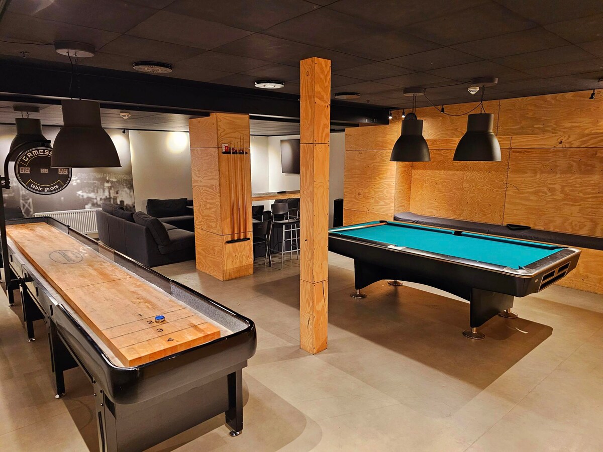 A spacious game room is equipped with a shuffleboard table and a billiards table, both featuring sleek designs. Comfortable seating is arranged nearby, illuminated by modern pendant lights. The walls are finished with smooth wood panels, contributing to a contemporary atmosphere.