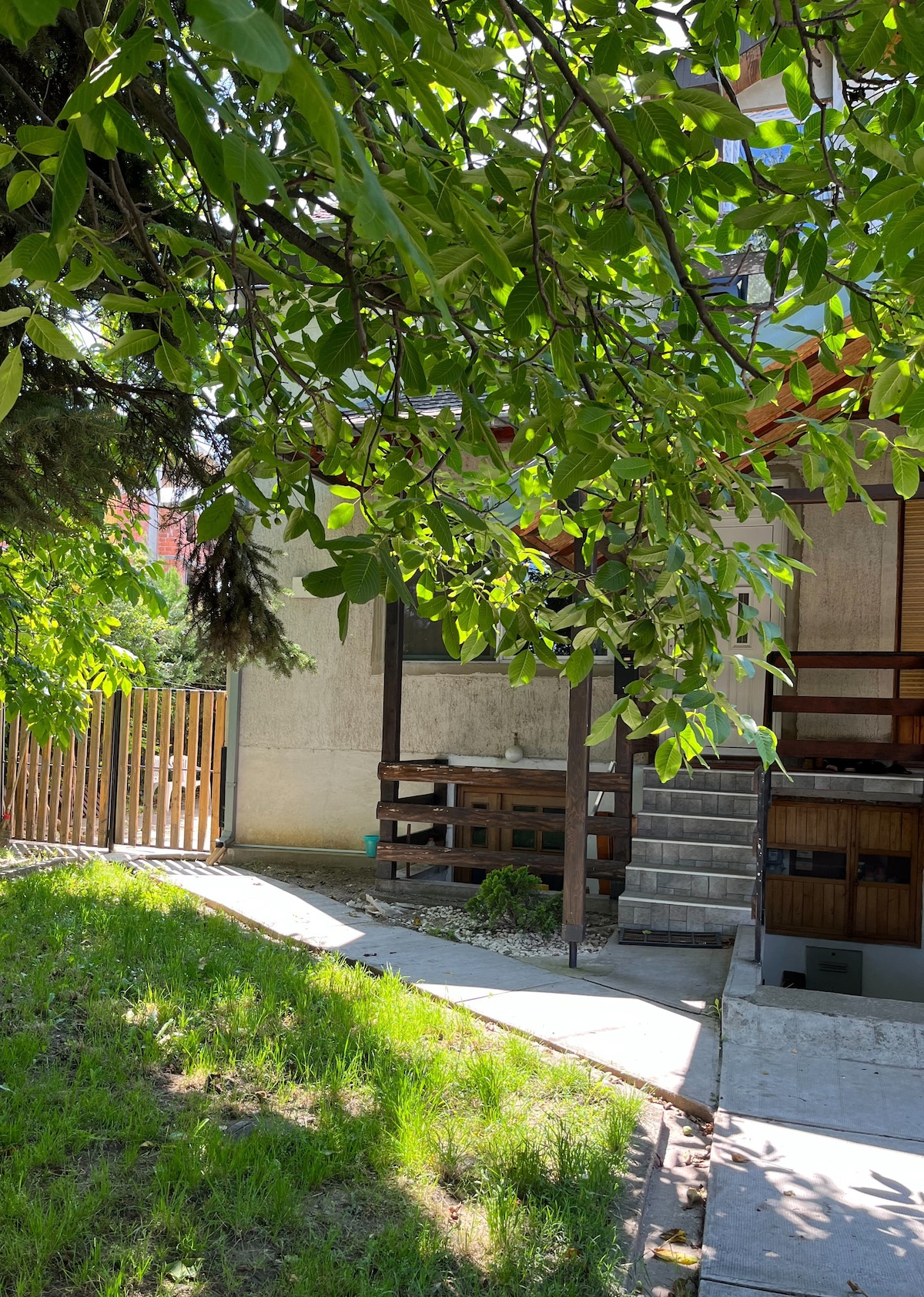 A pathway leads to a unique getaway surrounded by greenery, with a wooden porch supported by natural wood beams. Lush foliage partially frames the scene, while a wooden fence provides privacy. The area is designed to offer a tranquil outdoor experience.